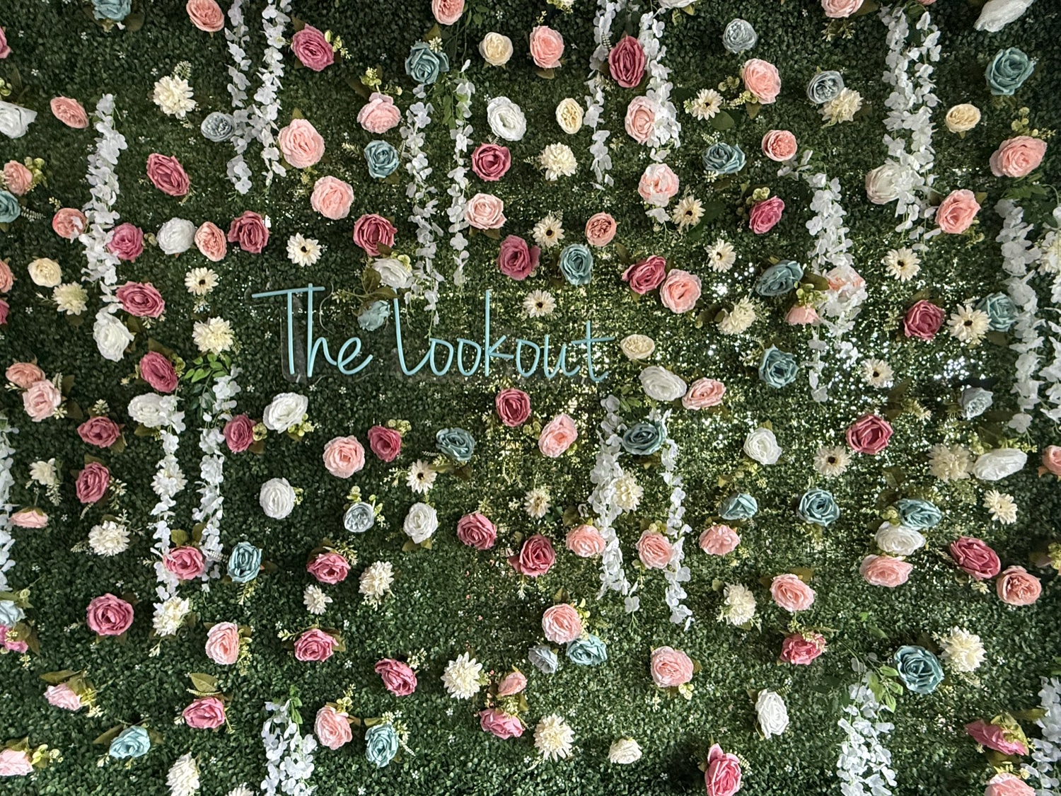 Teal neon sign reading The Lookout on a wall of flowers
